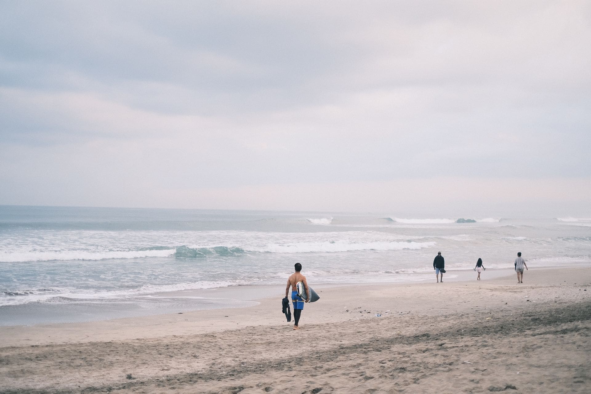 Una tranquila escena de playa en Bali, Indonesia, con surfistas dirigiéndose a las olas bajo un cielo nublado